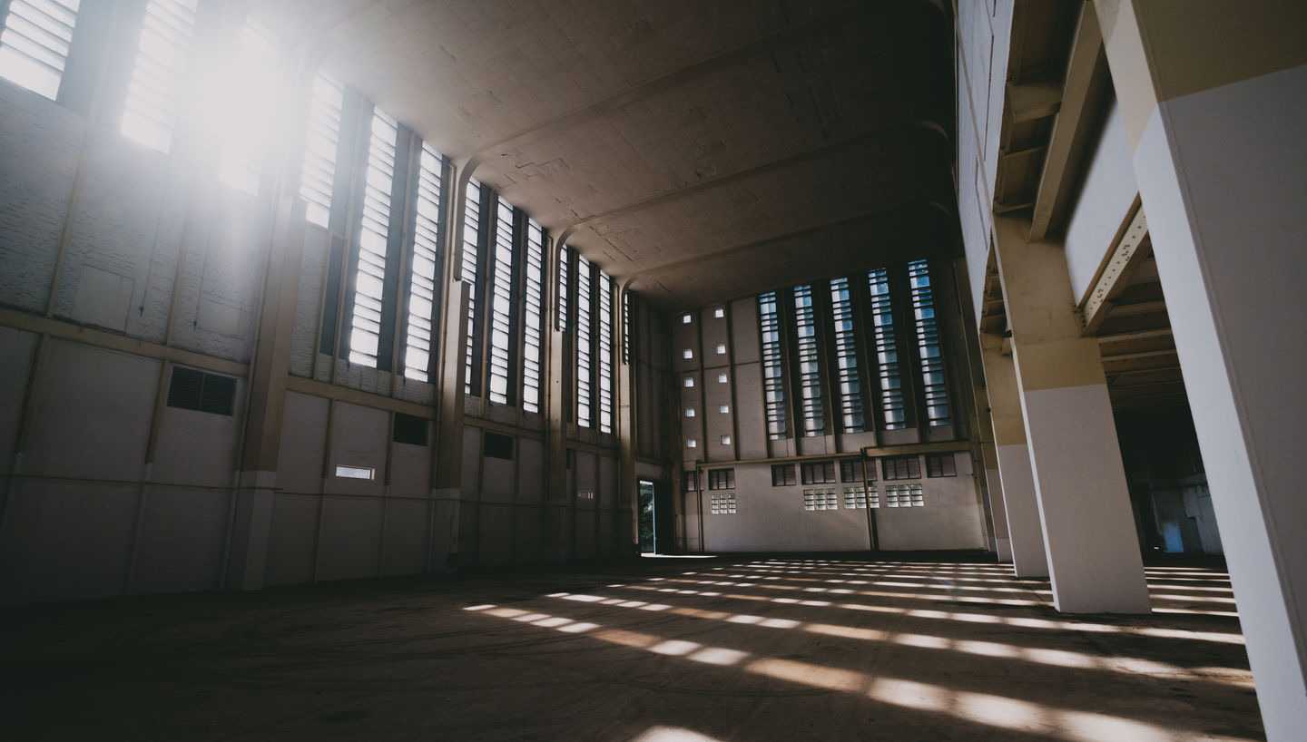 Heritage event hall interior — tall louvered windows, exposed concrete, sunlight streaming across the floor